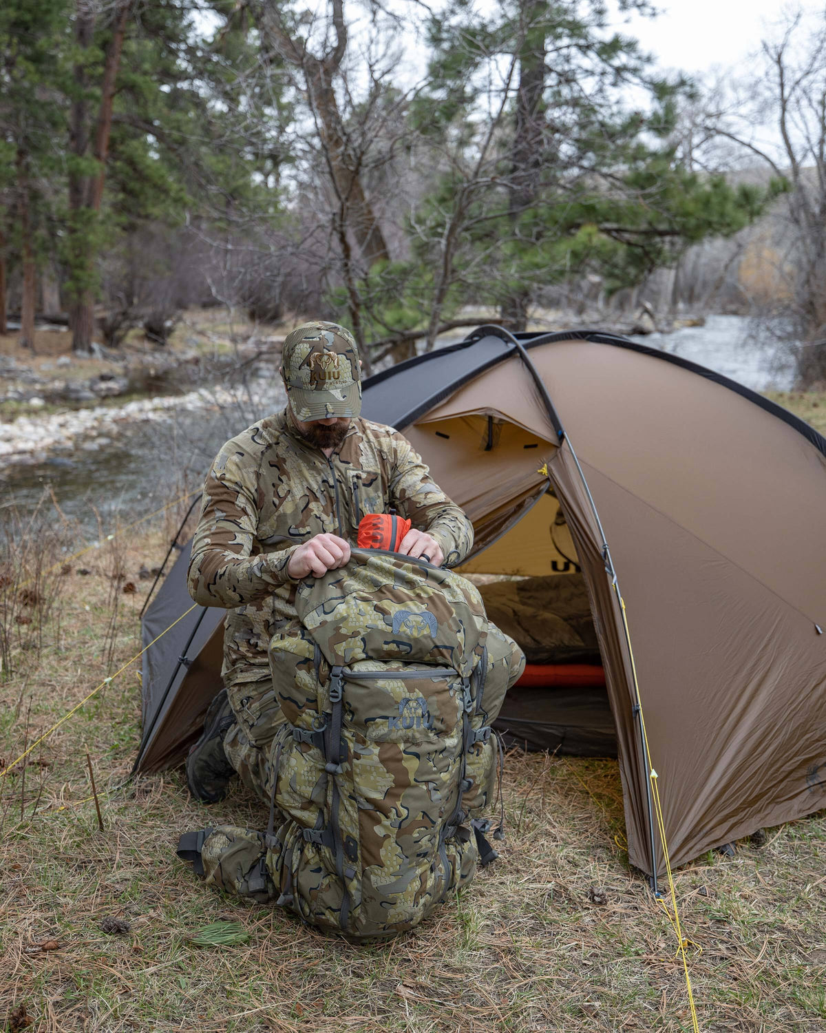 KUIU Mountain Star 3-Person tent in Gunmetal-Camel pitched beside a river in a pine forest