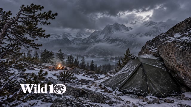 Taut small dark olive expedition tent on right sheltered behind a granite boulder, visible distant campfire safely on gravel beyond tent, windbent pine boughs and blowing snow, storm-gray dusk.